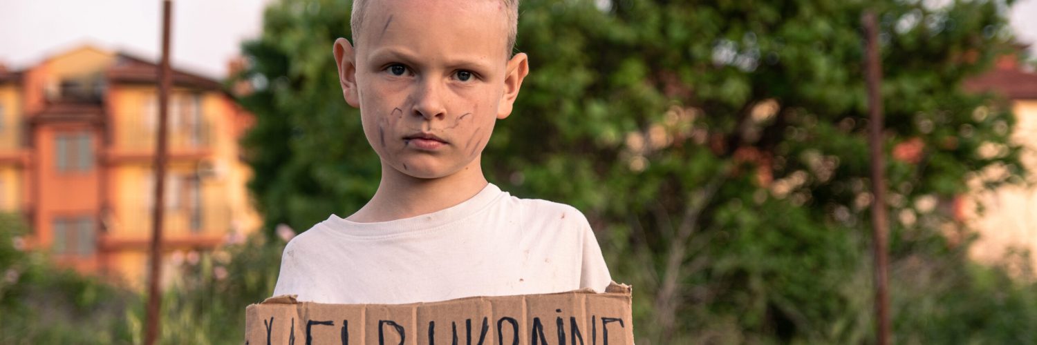 A boy with a grimy face and sad eyes holds a cardboard poster with the inscription HELP UKRAINE in English in his hands. A small boy of European appearance opposes military action.